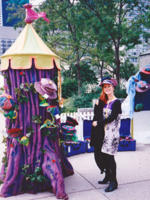 A woman wearing a Gwendolyne Hat beside the big purple display tree of Gwendolyne Hats