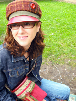 A close up of woman wearing a Abbey Road Cap in colours red and brown sitting on the grass with a Nathaniel shoulder bag