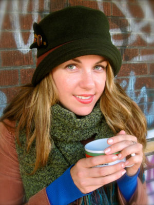 A woman sitting on a bench holding a cup of coffee and wearing a olive Corona Hat.