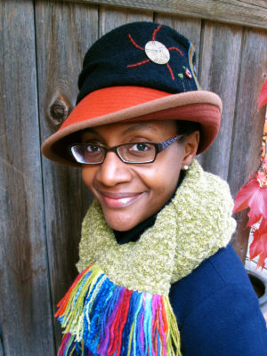 A close up of woman wearing a black and orange Harvest Moon hat design set against wood wall.