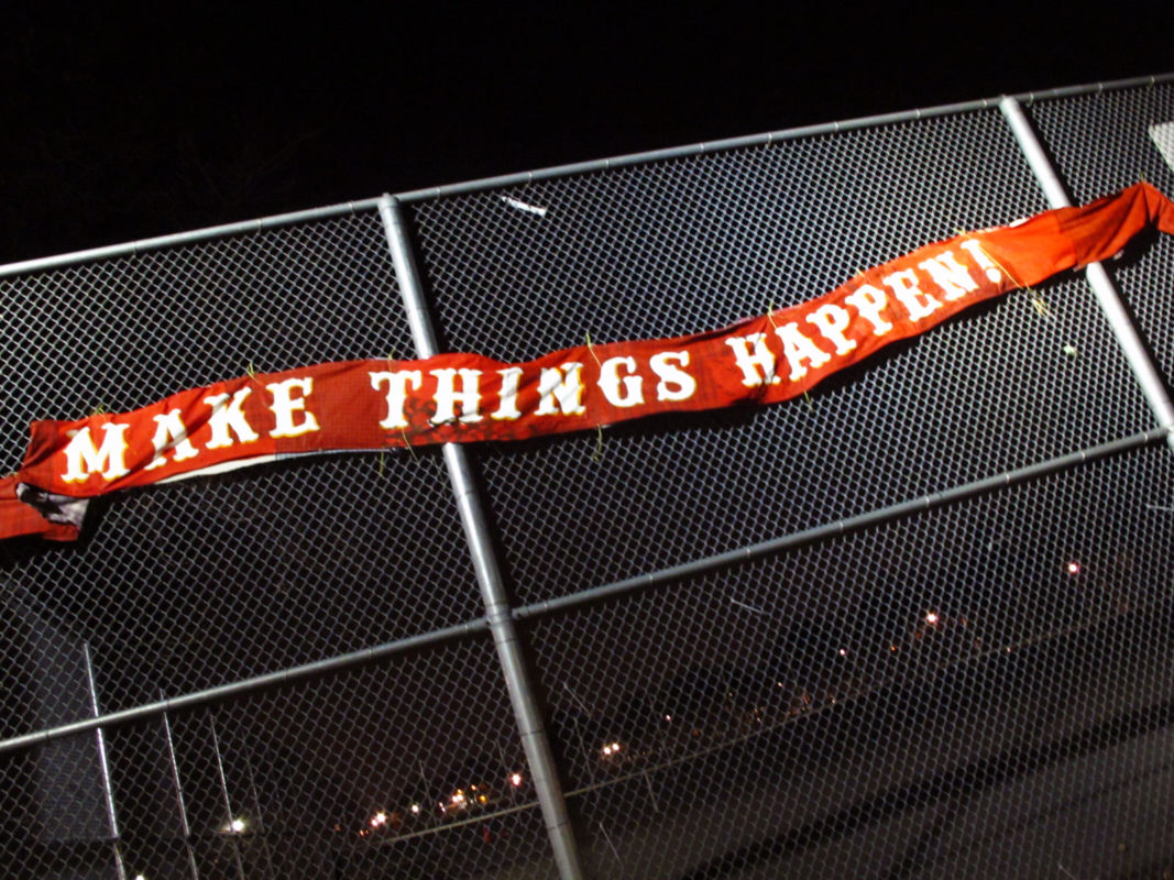 A red banner with the words Make things Happen in white block letters attached to a tennis fence photo shot in the night
