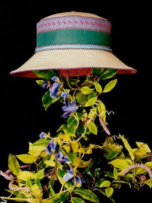 A blue and green summer wide rim Degas linen summer hat on a stand covered with leaves set against a black background